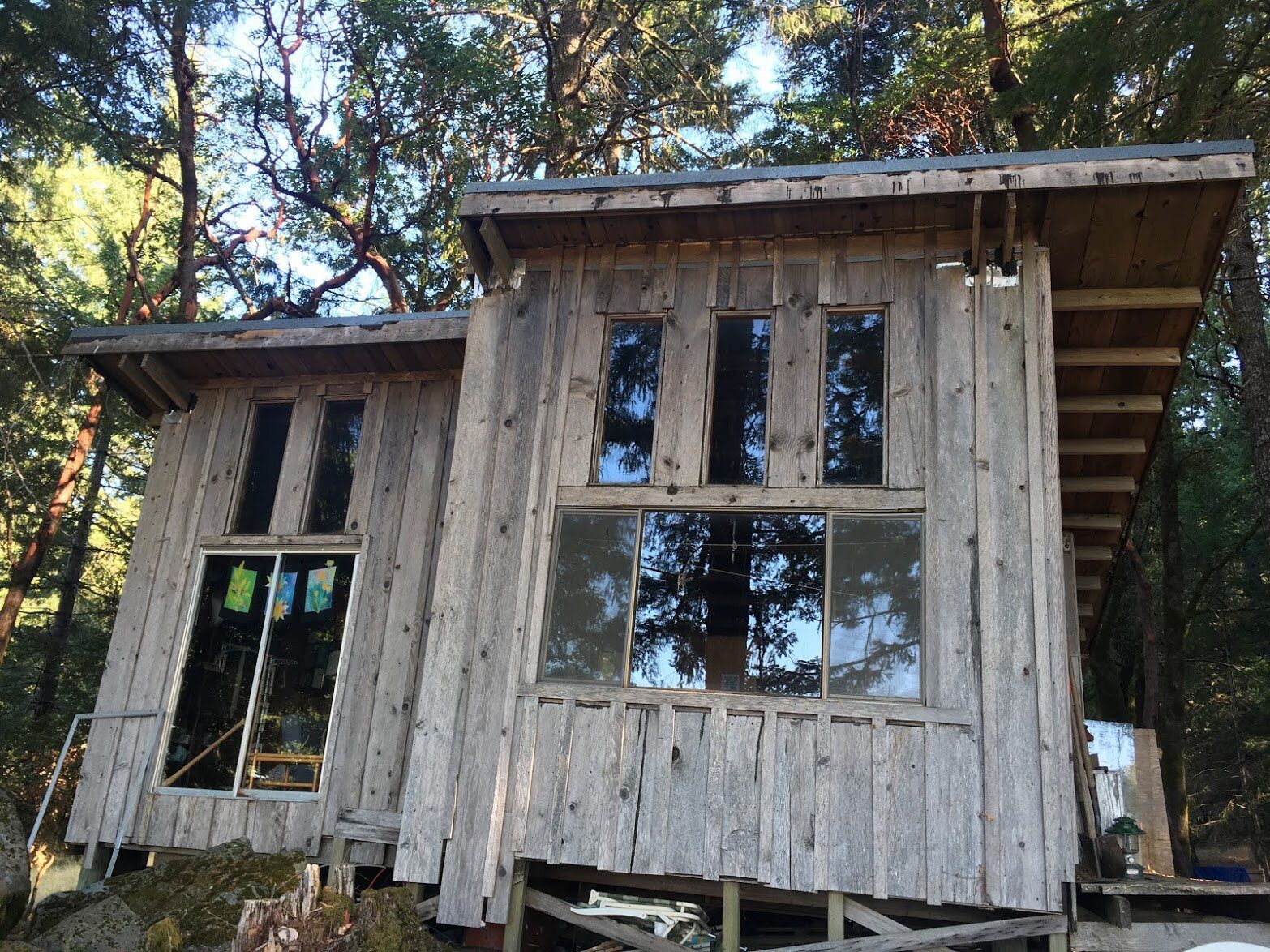 A two-story weathered board cabin with large windows, among trees.